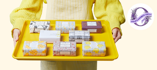 Two hands holding a yellow tray that has the eight bar soap from Beekman 1802's Sweet To Be Kind Bar Soap Set of 8 all lined up in three rows, with the model wearing a yellow sweater and striped apron on a cream colored background. 