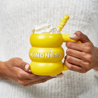 Up close shot of two hand holding Beekman 1802's Sunshine Ceramic Doughnut Mug, which has whipped cream and a yellow twisty candy coming out of the top, and the model's fluffy white sweater in the background. 
