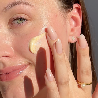 Up Close shot of side of models face while she is applying Beekman 1802's Milk Scrub Oat + Goat Milk Exfoliating Facial Cleaner to her cheek with her fingertips while looking at the camera.