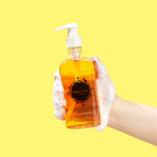 Sudsy hand holding a Clear bottle of Beekman 1802's Honeyed Grapefruit Hand & Body Wash with a white nozzle, on a yellow background.