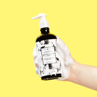 Sudsy hand holding black bottle of Beekman 1802's vanilla Absolute hand & body wash on a yellow background.