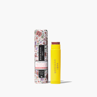 Yellow uncapped tube of Beekman 1802's Honeyed Grapefruit Sheer Tinted Lip Balm, standing next to packaging tube for lip balm thats covered in flowers, on a white background.