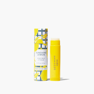 Yellow tube of Beekman 1802's Sunshine Lemon Lip Balm standing next to packaging covered with lemons, on a white background.