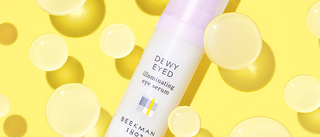 Bottle of Beekman 1802's Dewy Eyed Illuminating Eye Serum surrounded by round clear gel orbs on a yellow background.