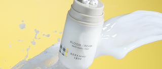 Bottle of Beekman 1802's Bloom Cream Daily Moisturizer mid-air, surrounded by a milky splash on a yellow background.