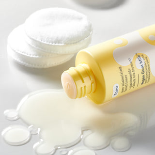 Yellow Bottle of Beekman 1802 Vegan Goat Milk Pore Minimizing Facial Toner laying flat with spilled toner underneath the toner, next to a stack of round cotton pads, all on a white background.