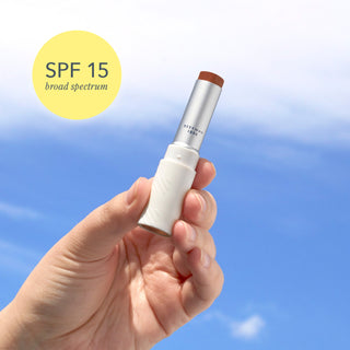 Upclose shot of hand holding Beekman 1802's Spicy Nudey SPF 15 Goat Milk Tinted Lip Cream with sky in the background and a yellow bubble to the right of the product that says "SPF 15 Broad Spectrum"