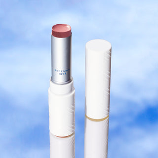 Tube of Beekman 1802's Rosy Posey SPF 15 Goat Milk Tinted Lip Cream opened with cap placed to the right of the tube on a blue sky background.