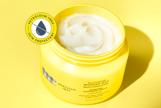Opened jar of Beekman 1802's Multipurpose Wonder Milk Buttery Botanical Jelly on a yellow background, with a callout on the top right that says "petroleum free," and "skin protectant,'" on a yellow background. 