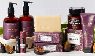 Products from Beekman 1802's Fig Leaf collection which includes the caddy set, lip balm, bar soaps, body cream, and hand cream all displayed next to each other, surrounded by sliced figs and leaves on a light grey tile background. 