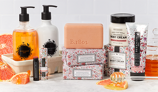 Products from Beekman 1802's Honeyed Grapefruit collection which includes the caddy set, lip balm, bar soap, and body creams, surrounded by slices of grapefruit and a honey stick spilling honey on a light grey tile background. 