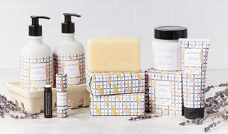 Products from Beekman 1802's Lavender Collection which includes the caddy set, lip balm, bar soap, body cream and hand cream all displayed next to each other, surrounded by lavender flowers on a light grey tile background. 