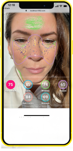 Cell phone photo of woman receiving her personalized results of the Skin Biome Analysis, with numerical results of texture, wrinkles, dark spots,  redness, hydration and skin biome indicated in color-specific areas on her face