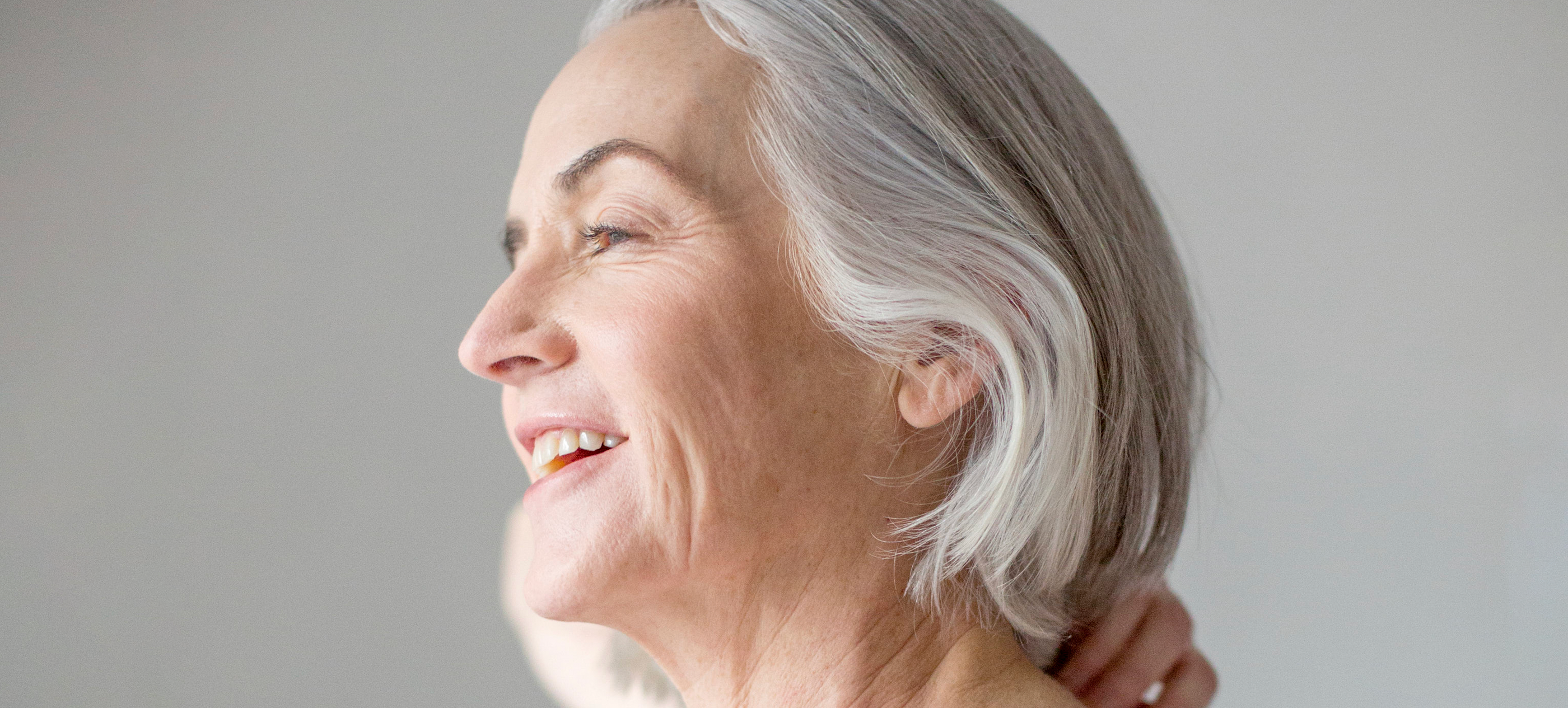 Head shot of mature model with grey short hair looking off to the side and smiling while holding the back of her neck.