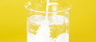 Shot of glass beaker filled with clear liquid, while milky white liquid is bring poured into the same beaker, on a yellow background.