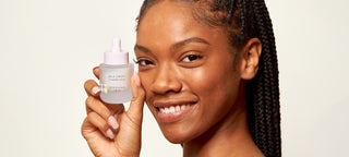 Close up head shot of model looking at the camera and smiling while holding a bottle of Beekman 1802's Milk Drop Ceramide Serum up to her face, on a white background.