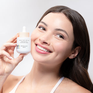 Image of model holding the Beekman 1802 Milk Tint SPF 43 Tinted Primer Serum next to her face while smiling and looking off to the side. 