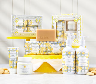 Collection shot of Beekman 1802's Almond Honey Cookie fragranced products all displayed next to each other on cake stands, surrounded by cookies and on a white background.