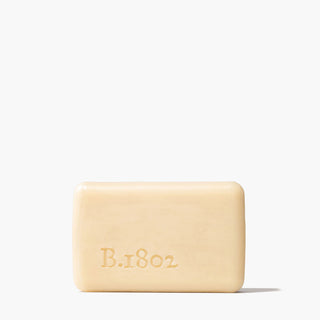 Unwrapped bar of Beekman 1802's Lilac Dream Goat Milk Soap, which has the word "B.1802" stamped on the bottom left of the soap, standing on a white background.
