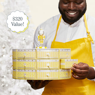Shot of male model wearing a yellow apron and holding Beekman 1802's 2023 Slice of Kindness Advent Calendar in one hand while looking down and pulling out on of the drawers with the other hand, with a white christmas tree in the background.