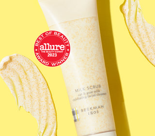 Tube of Beekman 1802's Milk Scrub Oat & Goat milk Exfoliating Cleanser laying flat on a yellow background, surrounded by the product smears and next to a red seal that says "Allure 2023 Best Of Beauty Award Winner."