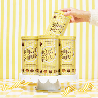 4 yellow tins of Beekman 1802's Ultra Premium Goat Poop Chocolates on a white cake stand surrounded by yellow ribbons, with a hand coming out of the right side and pulling out one of the tins, on a yellow and white striped background. 