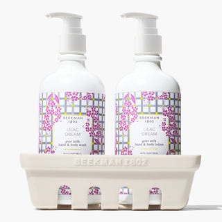 Up close shot of Beekman 1802's Lilac Dream Caddy Set, which includes both white bottles with white nozzles of the lilac dream Lotion on the right, and lilac dream hand & body wash on the left, both sitting inside a ceramic caddy that says "beekman 1802" on the front, on a white background.