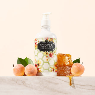 Bottle of Beekman 1802's Apricot & Honey Tea Goat Milk Lotion, surrounded by apricots and dripping honey on a marble counter, on a cream colored background. 