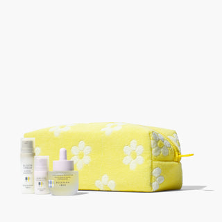 Beekman 1802's Tighten Up Skincare Set which includes a mini bloom cream daily moisturizer, a mini dewy eyed eye serum, and a full size collagen booster, and are all lined up in front of a yellow dopp bag with white flowers, on a white background.