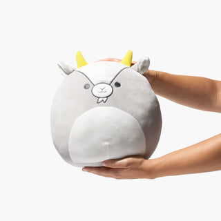 Shot of hand holding up Beekman 1802's Goatie Squishmallow which is in the shape of a cartoon goat with yellow horns for scale, on a white background.