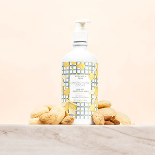 White bottle of Beekman 1802's almond honey cookie lotion with a blue and grey gingham label, surrounded by almonds and cookies and standing on a marble counter, on a cream colored background.