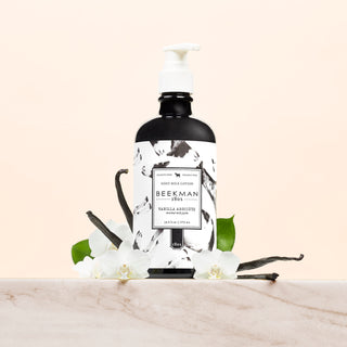 Black Bottle of Beekman 1802's Vanilla Absolute Goat Milk Lotion, with a white pump and white label on it, standing on top of vanilla beans and white flowers on a marble counter, on a cream colored background.