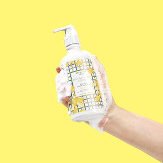 Sudsy hand holding a bottle of Beekman 1802's Almond Honey Cookie Hand & Body Wash with a white nozzle, on a yellow background.