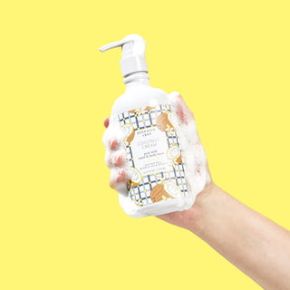 Sudsy hand holding a bottle of Beekman 1802's Coconut Cream Hand & Body Wash with a white nozzle, on a yellow background.
