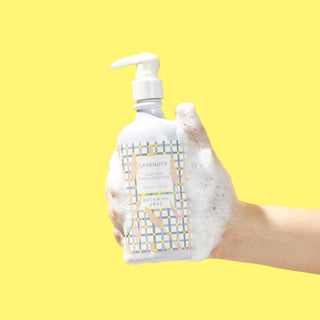 Sudsy hand holding a bottle of Beekman 1802's Lavender Hand & Body Wash with a white nozzle, on a yellow background.