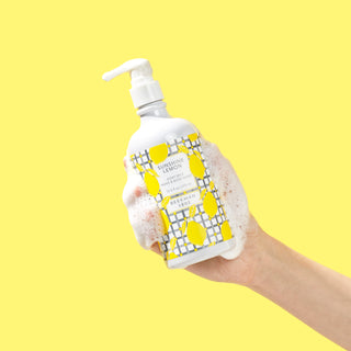 Sudsy hand holding a bottle of Beekman 1802's Sunshine Lemon Hand & Body Wash with a white nozzle, on a yellow background.