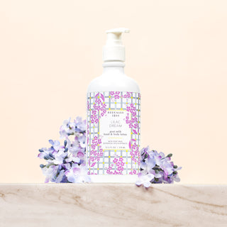 white Bottle of Beekman 1802's Lilac Dream Lotion with with gingham label designed with lilac flowers and a white nozzle on top, surrounded by purple lilac flowers and standing on a marble countertop, on a cream colored background.