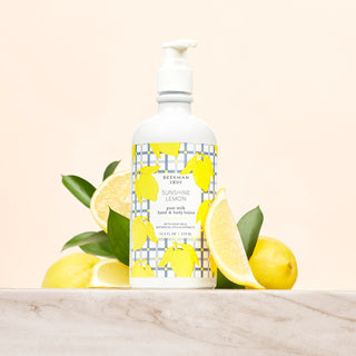Bottle of Beekman 1802's Sunshine lemon lotion standing on a marble countertop surrounded by leaves and sliced lemons on a cream colored background. 