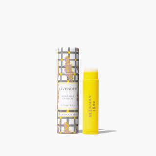 Yellow uncapped tube of Beekman 1802's Lavender Lip Balm standing next to the packaging tube for the lip balm that is designed with lavender flowers and gingham pattern, on a white background. 