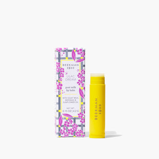 Yellow tube of Beekman 1802's uncapped Lilac Dream Lip Balm with the words "Beekman 1802" stamped on the lip balm, standing to the right of the packaging box for the lip balm that is designed with blue and grey gingham and covered in lilac flowers design, on a white background.