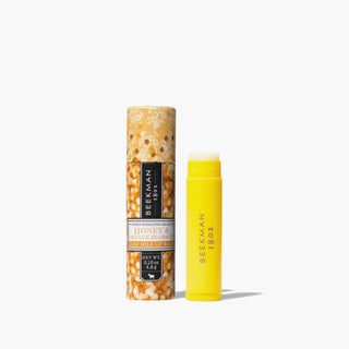 Yellow uncapped tube of Beekman 1802's Honey & Orange Blossom Lip Balm, standing next to orange packaging tube that is designed with flowers, on a white background. 