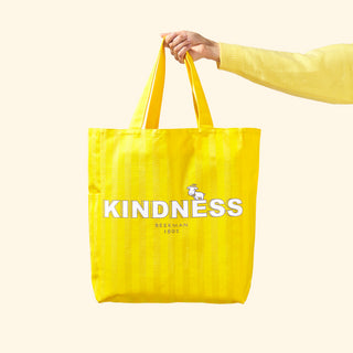 Image of arm wearing a yellow sweater and holding up beekman 1802's 2023 Sunshine Yellow Holiday Goat Getter tote bag which says, "Kindness, Beekman 1802," on the front, with a cartoon goatie on top of the letters, on a eggshell colored background.