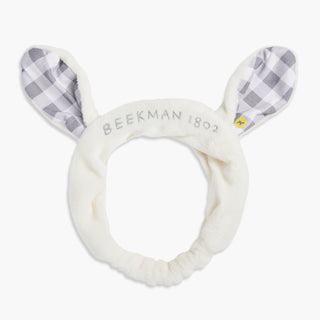 Beekman 1802's Goat Ears Headband, with white and grey gingham pattern in the ears and with the words "Beekman 1802" written across the front of the headband, on a gray backgorund.