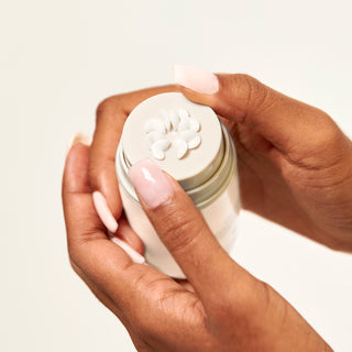 Bottle of Beekman 1802's Bloom Cream Daily Moisturizer being held with both hands while product is being pumped out at the top on a white background.
