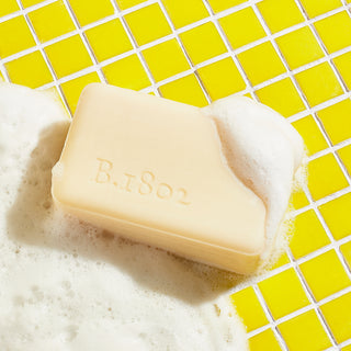 Sudsy Beekman 1802 almond Honey cookie goat milk palm-sized soap on yellow bathroom tile, next to sudsy water.