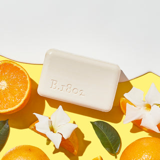 Unwrapped Honey & Orange Blossom goat milk bar soap on a yellow backgorund, surrounded by orange slices and white flowers, and a goat milk spill coming from the top.