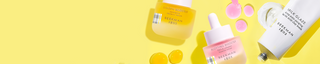 Bottle of Beekman 1802's Blotting Booster PHA & Calamine Blemish Serum, Golden Booster Vitamin C & amla berry serum, and Tube of Beekman 1802's Milk Glaze Clay Mask on a yellow background, surrounded by booster serum droplets and milk glaze product smear behind the products.