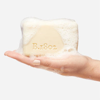 Up close shot of Hand holding sudsy bar of Beekman 1802's Garden of Gardenia Goat Milk Soap on a grey background