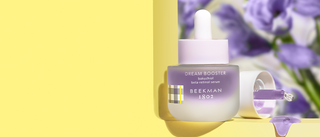 Bottle of Beekman 1802's Dream Booster Bakuchiol Beta-Retinol Serum on a yellow window sill, next to a serum dropper with the purple serum dripping onto the sill, with purple flowers in the background.
