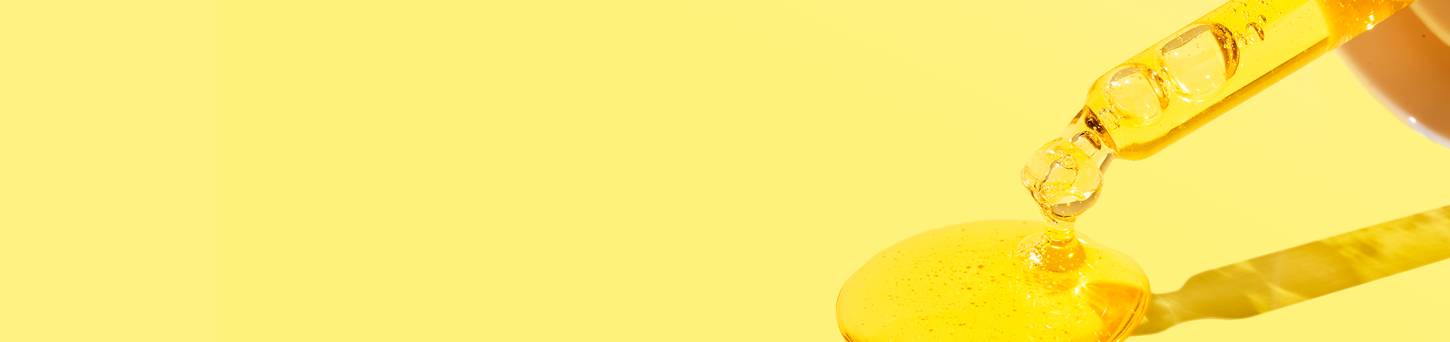 Closeup shot of serum dropper from Beekman 1802's Golden Booster Vitamin c & amla berry Brightening Serum, dropping orange serum onto the yellow ground to the far right of the screen